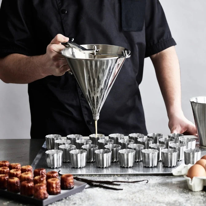 A stainless steel automatic funnel, also known as a sauce gun, being held by a chef dispensing liquid into stainless steel molds.  It has a tapered shape with a spout and a trigger mechanism, with no stand, being hand held.