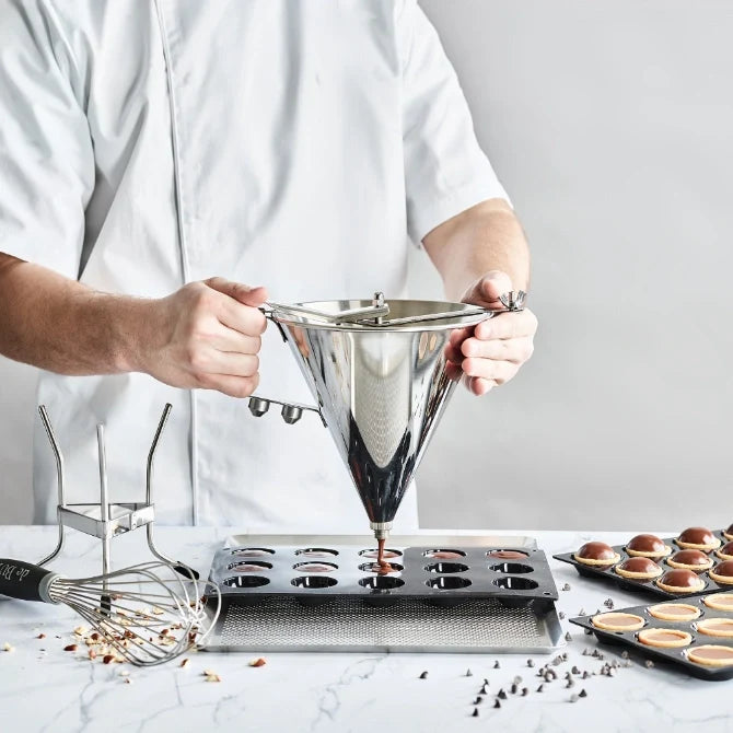 A stainless steel automatic funnel, also known as a sauce gun, being held by a chef dispensing liquid. It has a tapered shape with a spout and a trigger mechanism, with no stand, being hand held.