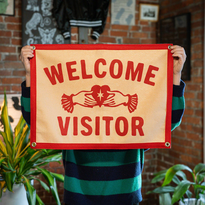 Person holding a 'Welcome Visitor' sign with a brick wall and plants in the background